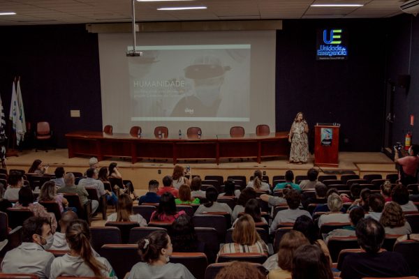 [:pt]Em um auditório estão diversas pessoas sentadas vendo uma tela com o banner do curta-metragem Humanidade. No palco, há mesas e cadeiras. Sobre o palco está uma mulher loira, de vestido longo, falando ao microfone.[:]