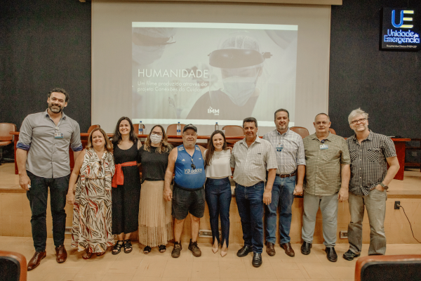 [:pt]Há 10 pessoas na foto, sendo 4 mulheres e 6 homens. Atrás deles, há um telão com a imagem de uma médica com paramentos de covid-19 e ao dela o título "Humanidade".[:]