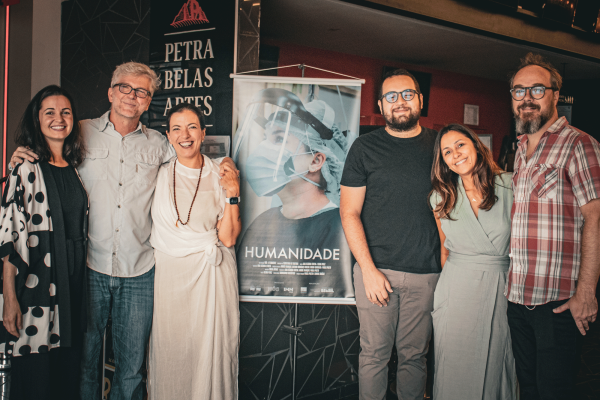 [:pt]Na foto há um banner do curta-metragem Humanidade ao centro. O banner tem a imagem de uma médica durante a pandemia e o título do curta-metragem. Do lado direito, estão 2 mulheres e um homem. Do lado esquerdo estão 2 homens e uma mulher. Todos profissionais que realizaram o documentário.[:]